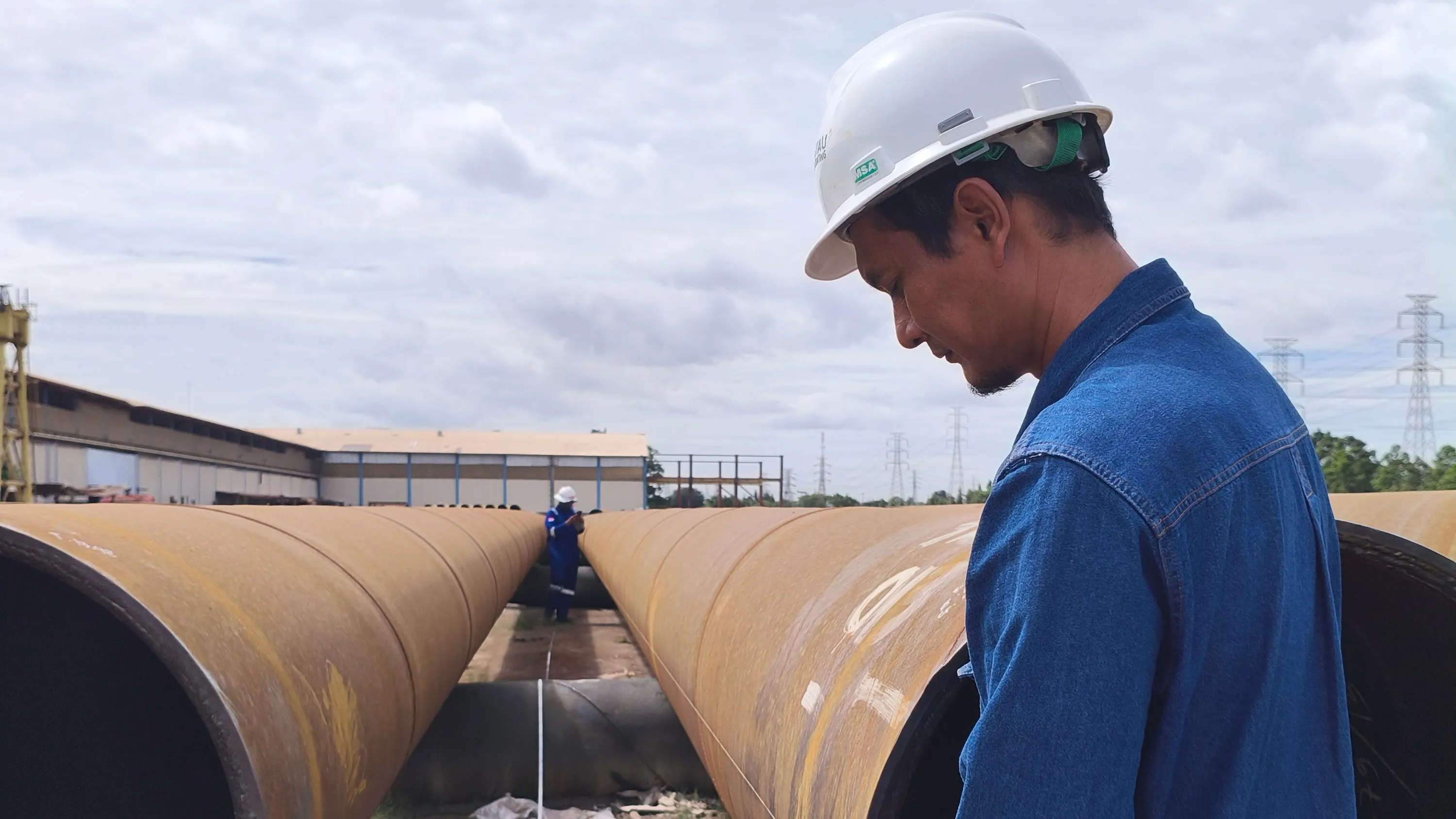 Two workers inspecting large-diameter pipe in a yard, one taking a measurement along the pipe run.
