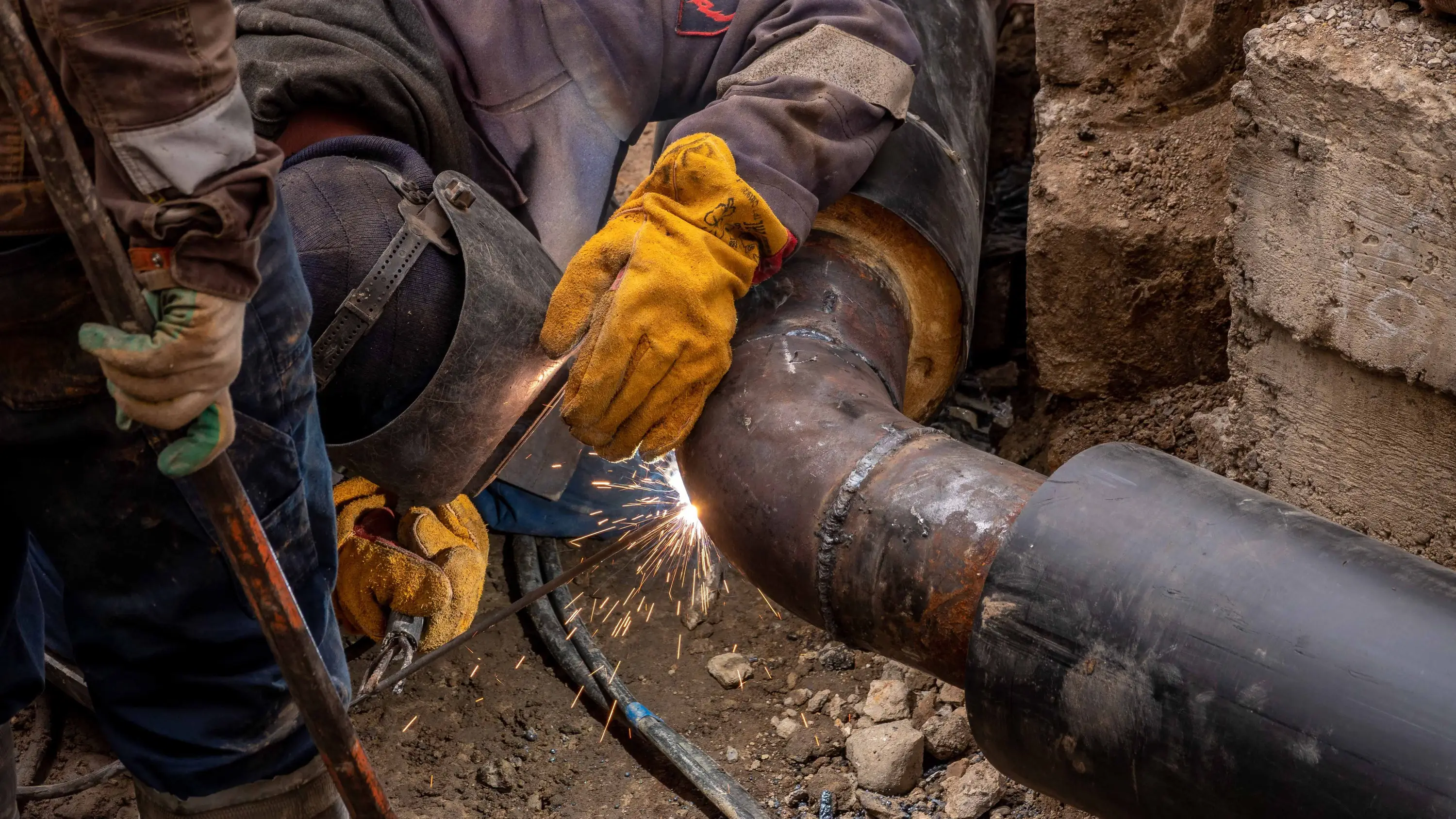 A welder joining a section of coated steel pipe in an open trench, with a second worker assisting.