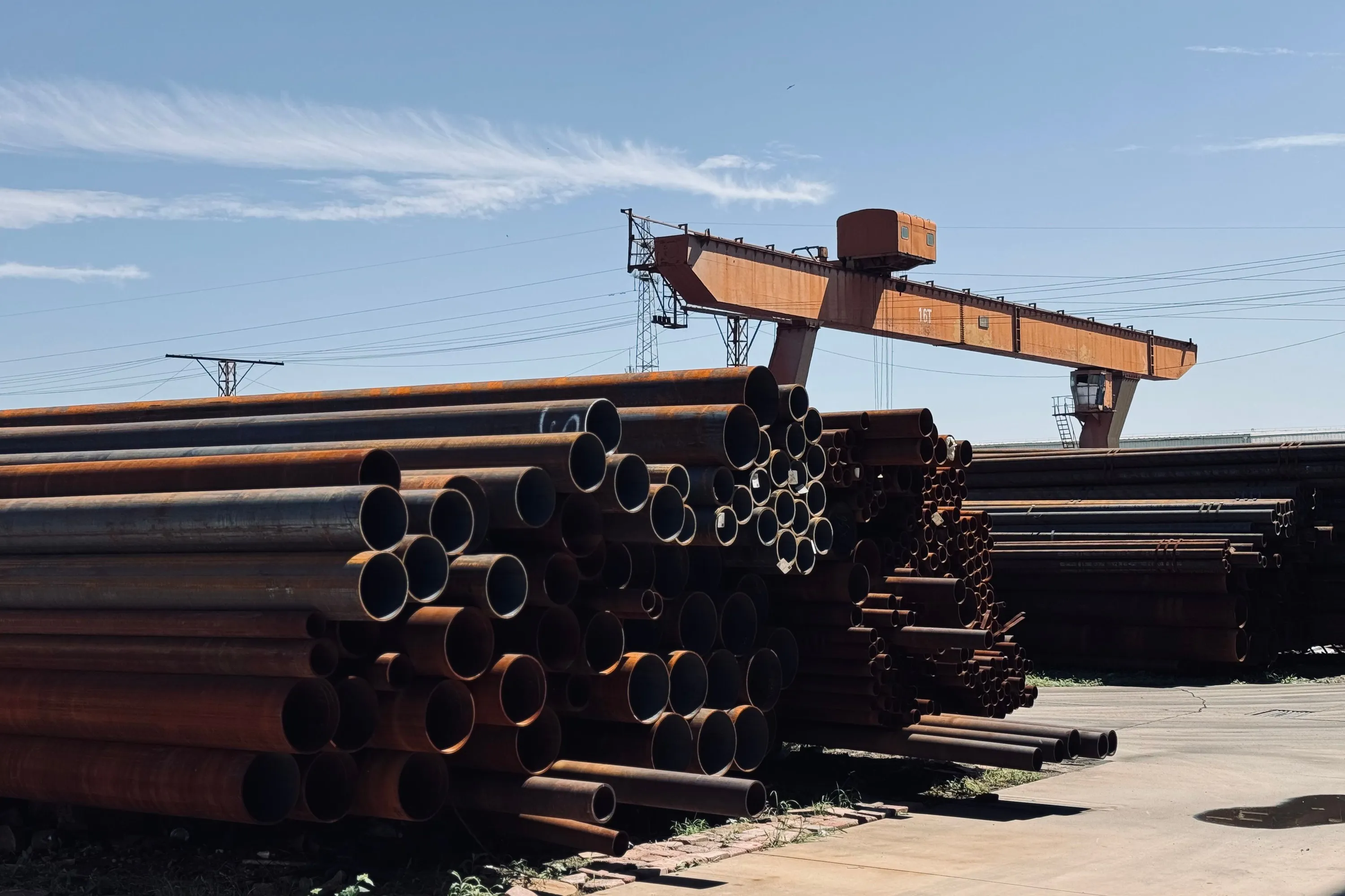 Large-diameter steel line pipe stacked in an outdoor pipe yard beneath a gantry crane.